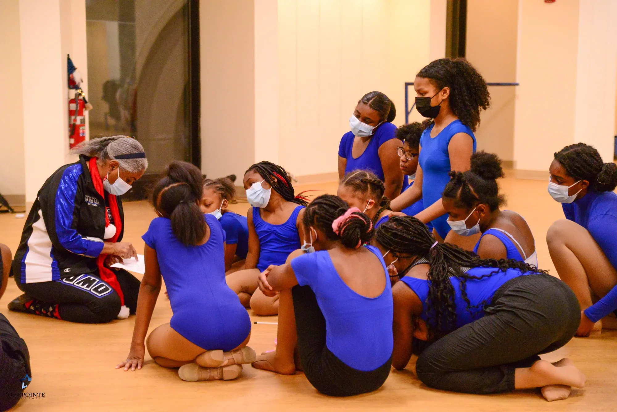Debra White-Hunt kneels on the studio floor with a circle of young dancers in blue leotards.